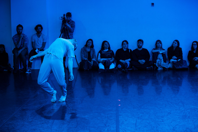 Alex bends his torso backwards wearing sweats. Audience members sit against a wall to watch.The lighting is a deep blue.